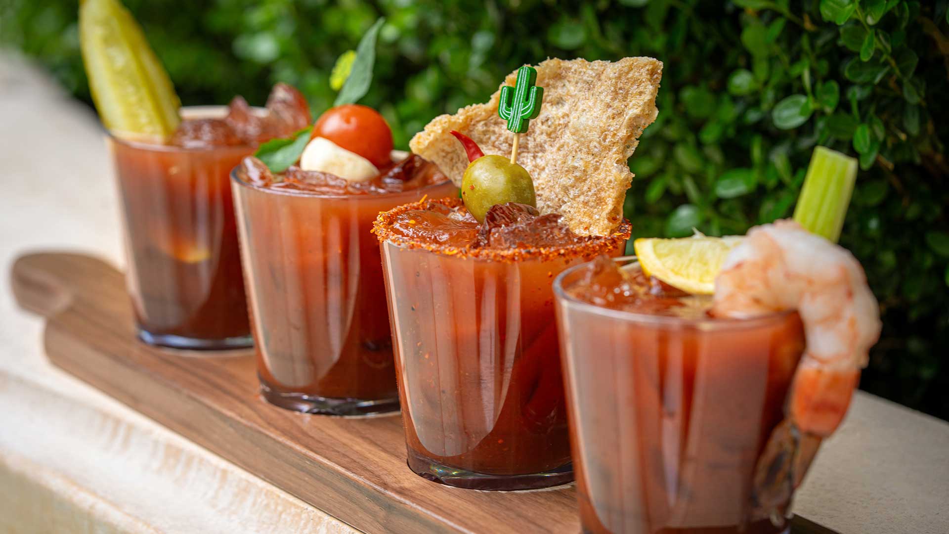 Four bloody mary drinks sit on a wood board in front of a green shrub.