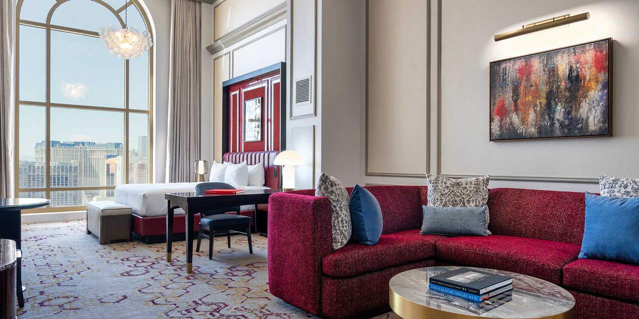 A hotel room with a king bed, desk with a chair, curved red couch, and a gold coffee table. 