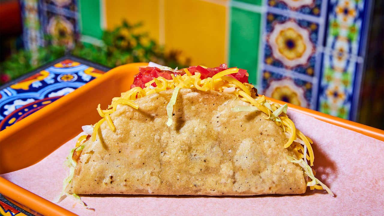 Large hard-shell taco with cheese and tomatoes on a orange tray in front of a colorful wall.