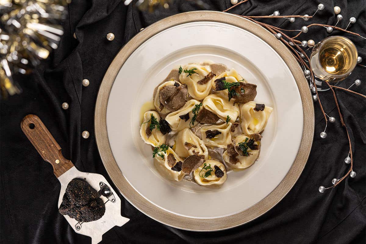 Cappellacci pasta on a plate covered in Castelrosso sauce and shaved black truffle sitting on a black table top along a truffle shaving tool, a stemmed glass filled with a drink and some decorative branches