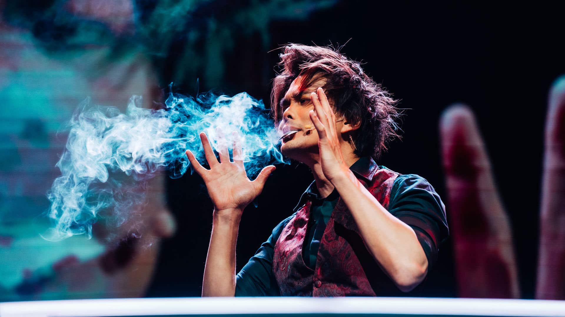 Shin Lim framing his face with this hands with a card and smoke coming out of his mouth.