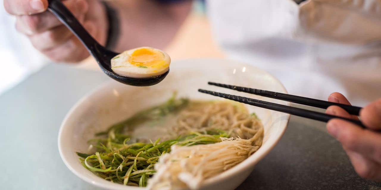 Ramen soup in a bowl with someone holding a hardboiled egg in a spoon and chopsticks over the bowl.