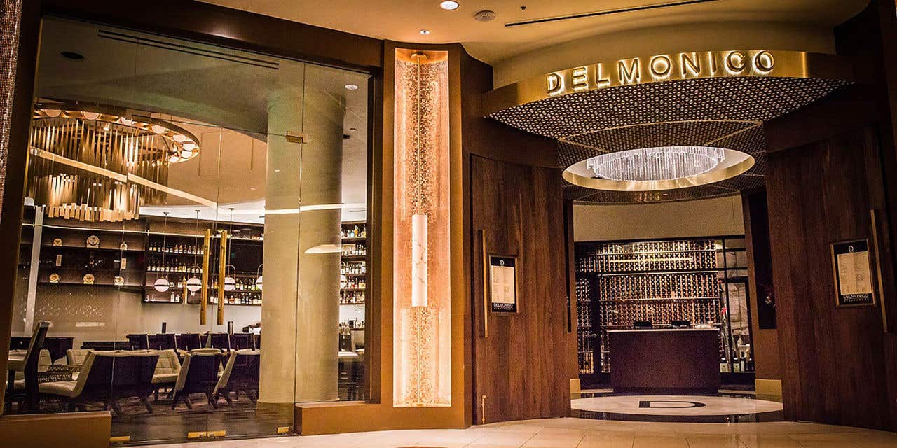 A glass fronted restaurant in warm wood tones with gold accents. Double doors are open under the name Delmonico.
