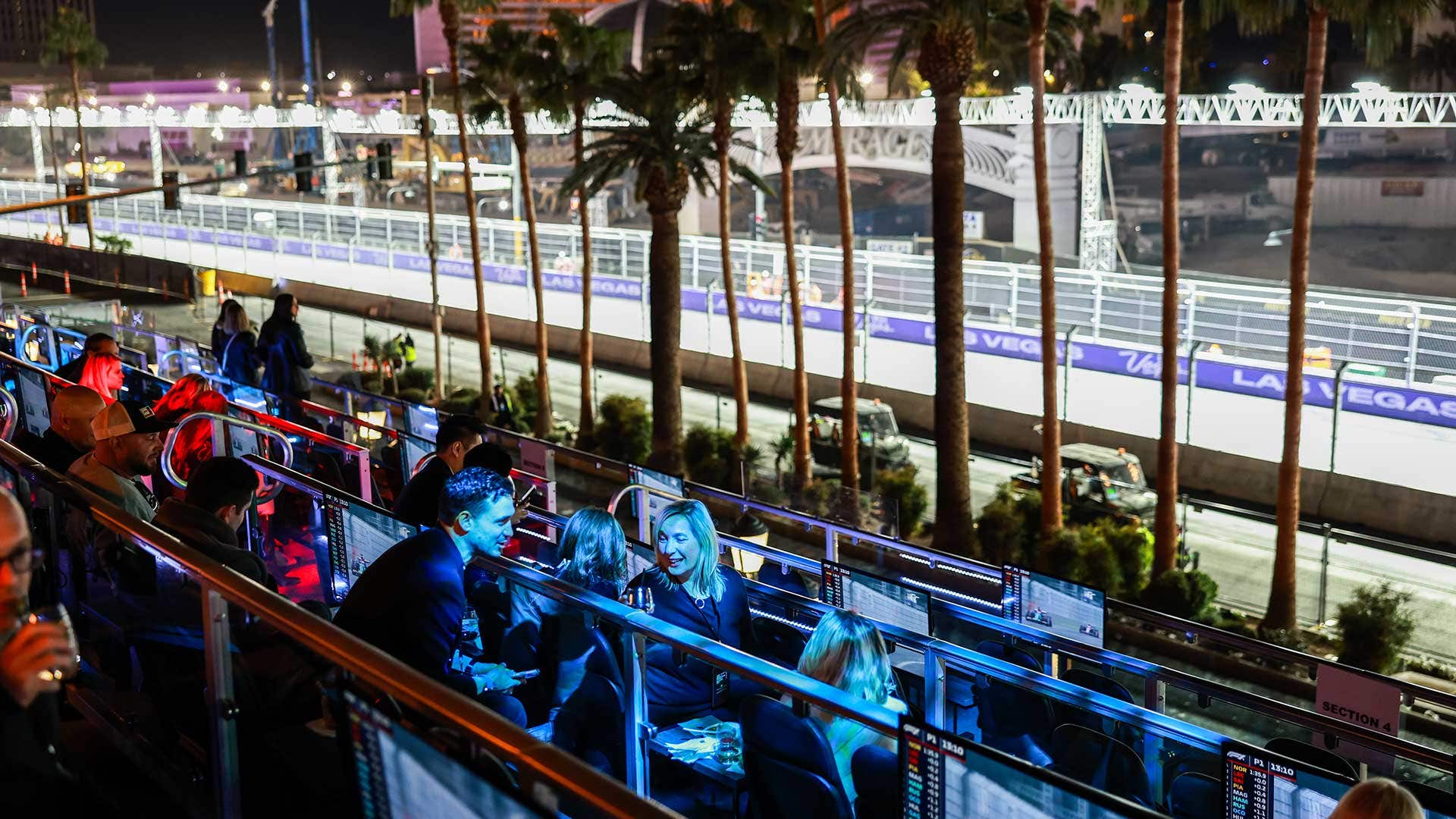 Racegoers chatting amongst themselves under a blue light while sitting on a viewing platform, with an illuminated racetrack in the background