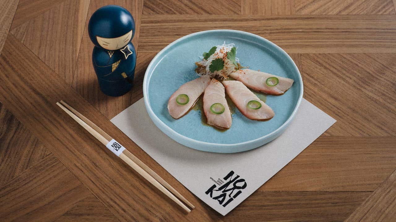 Plate of yellowtail sashimi with green pepper slices on a wooden table next to a decorative doll and chopsticks.