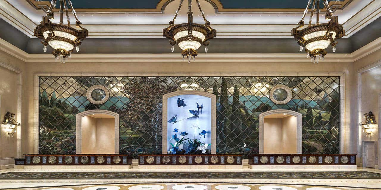 A spacious hotel lobby with three chandeliers and a long check-in desk, with a display of large blue butterflies behind it.