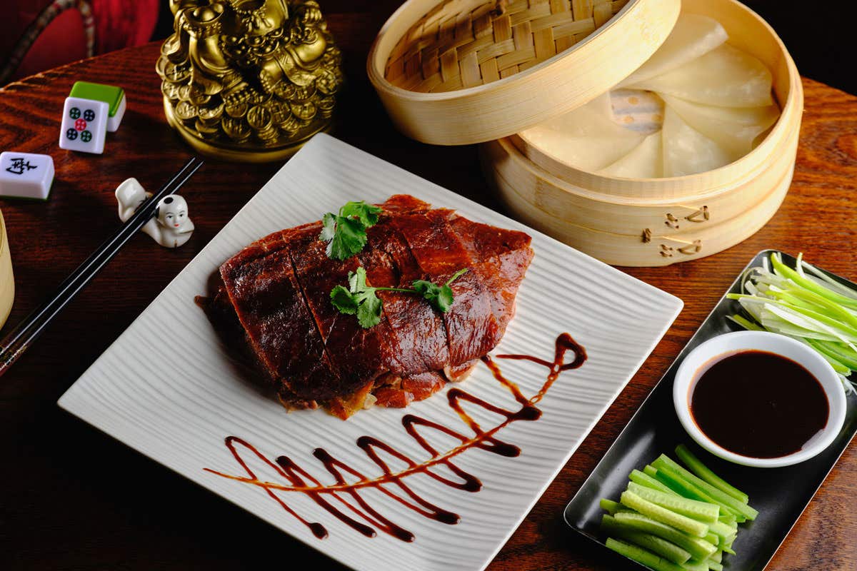 A plate with Peking duck pieces with a sauce decoration, placed next to a plate of garnishes and sauce, as well as a steam basket of pancakes to wrap the duck in