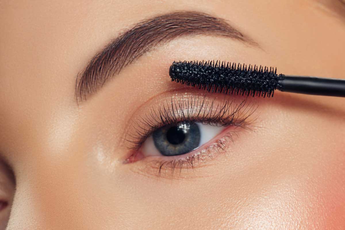 A close up of a woman’s blue eye with a manicured eyebrow and eyelashes, and a mascara wand.