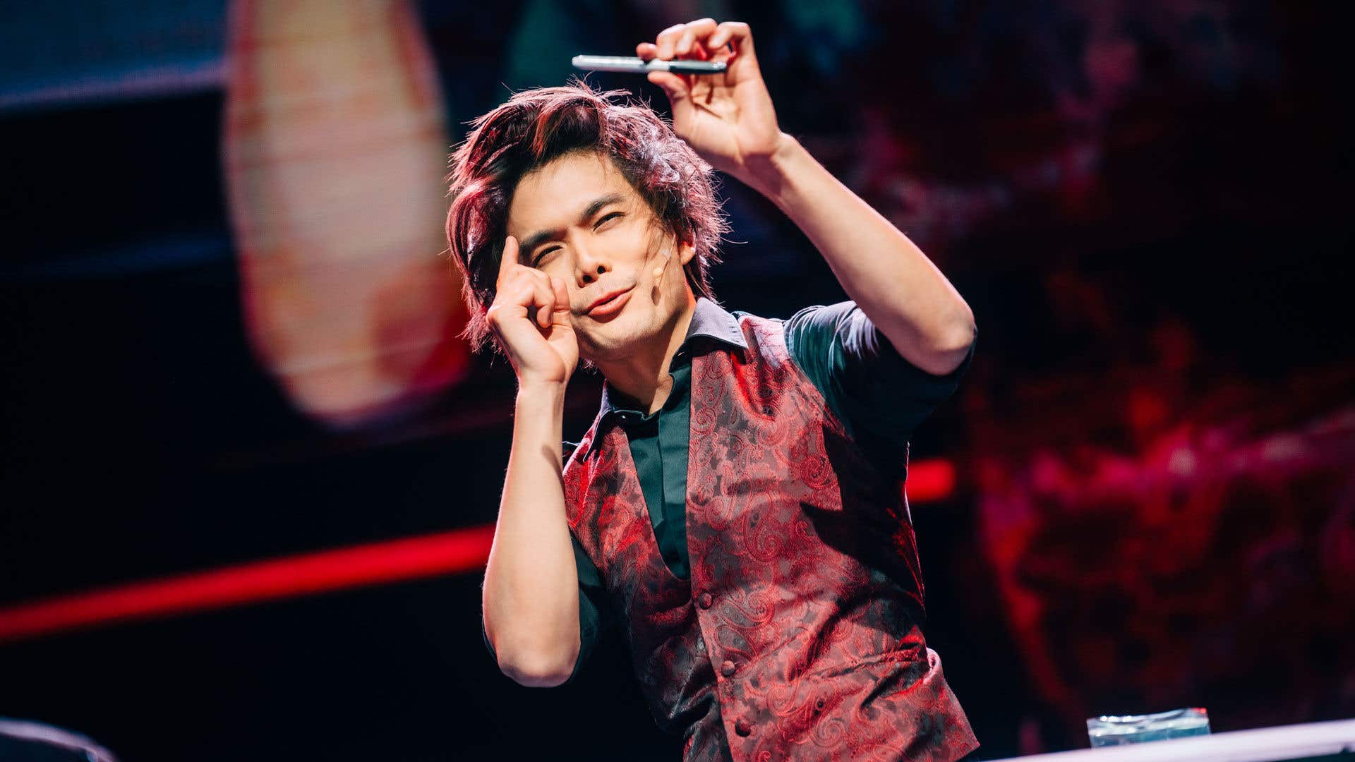 Shin Lim in a black shirt and red vest holding a pen in the air with one hand and pointing at it with his other hand. 