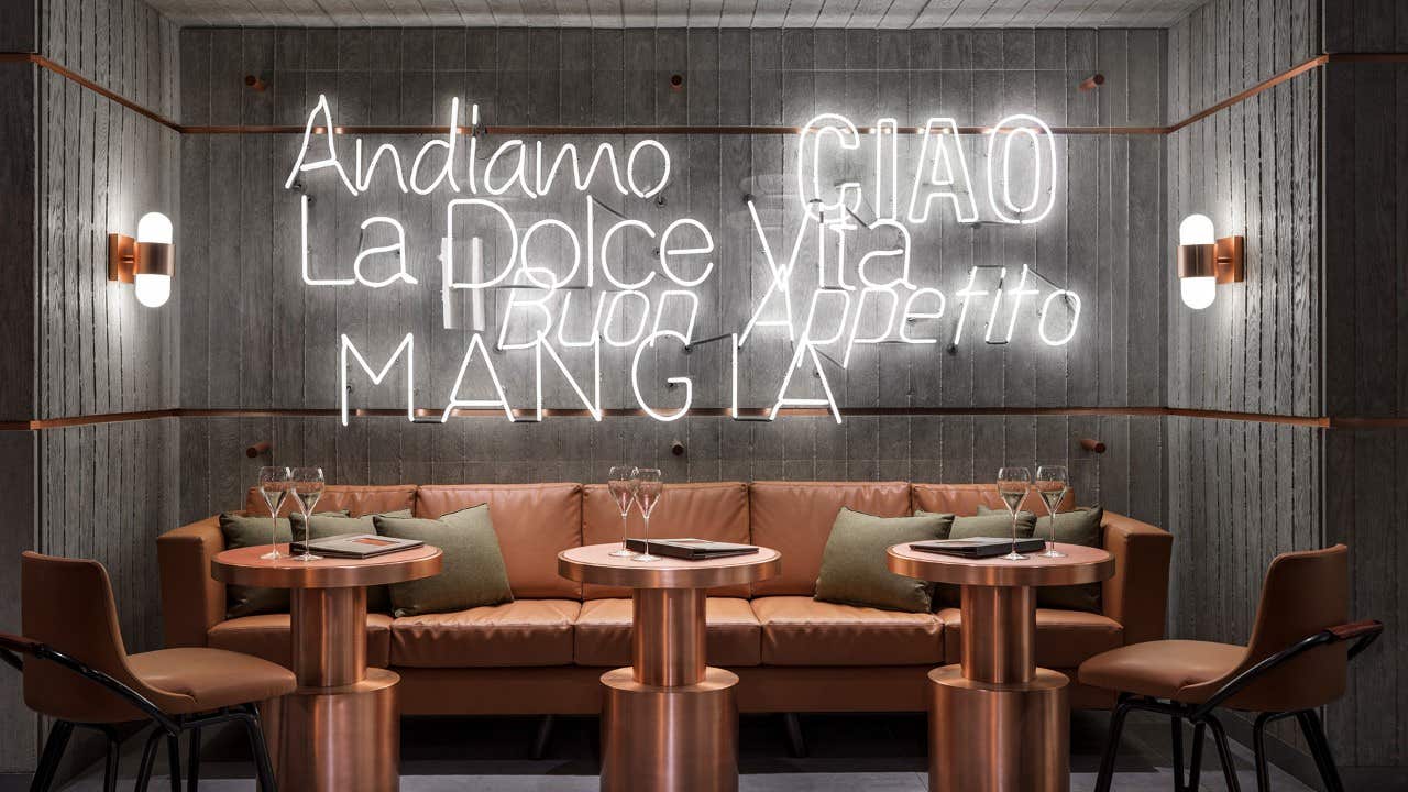 Modern dining area with a brown leather sofa, marble tables, and neon signs in Italian on a concrete wall.